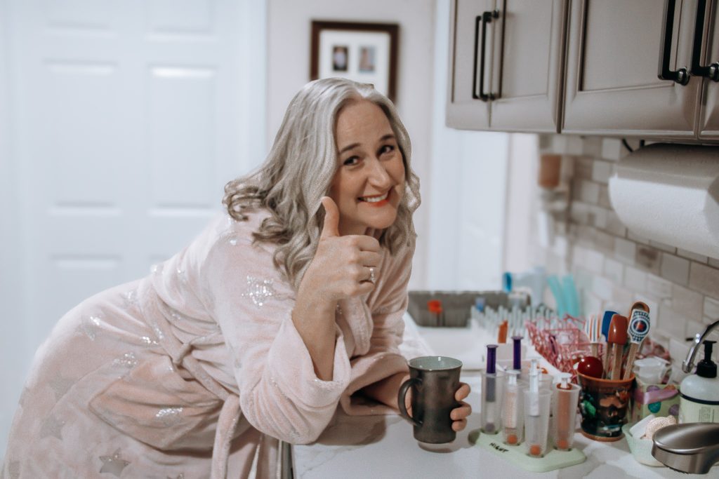 Jennifer Swann in a comfortable bathrobe holds a mug, giving a "thumbs up."