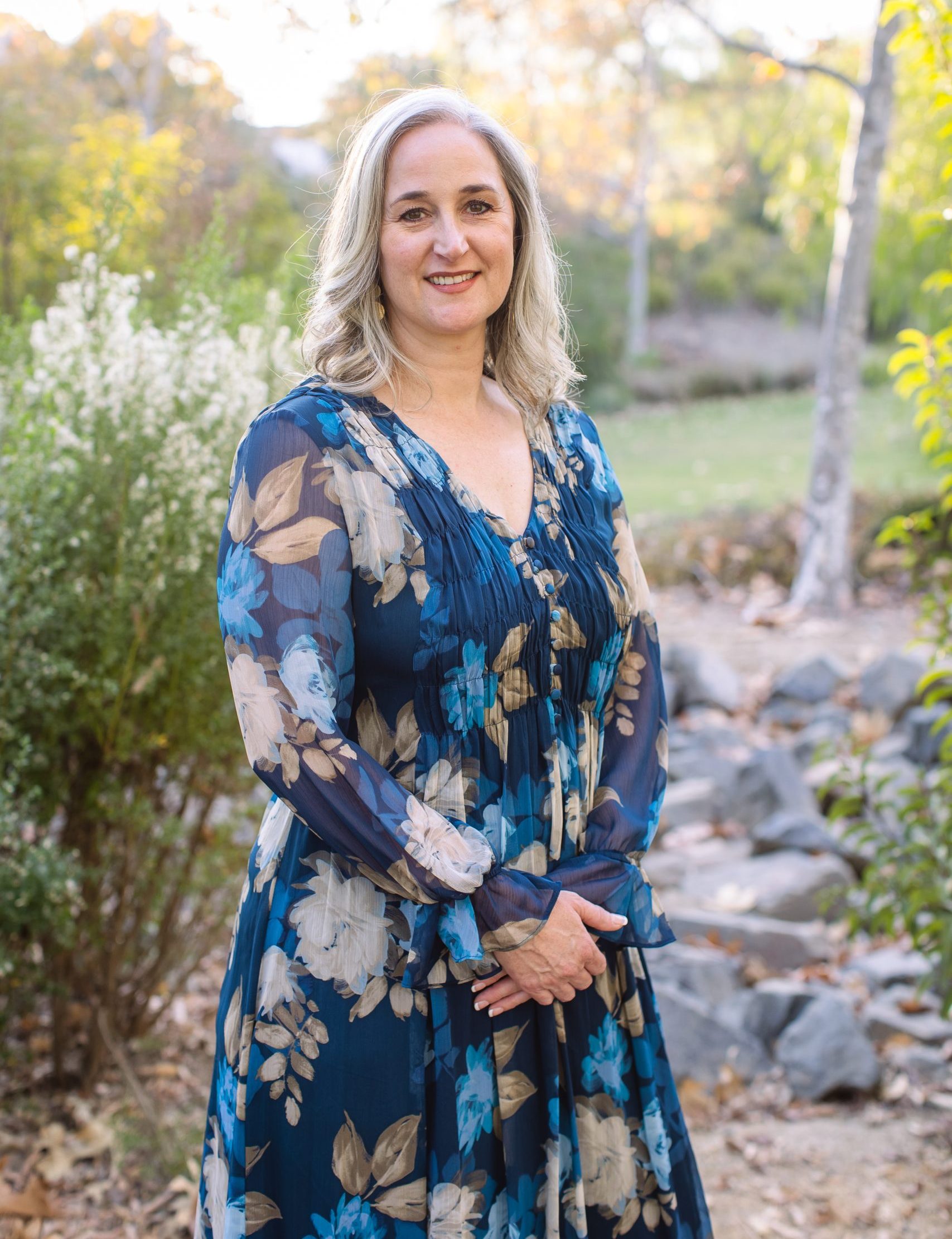 Jennifer stands in a blue dress with a backdrop of rocks, flowers, and trees on a sunny day,
