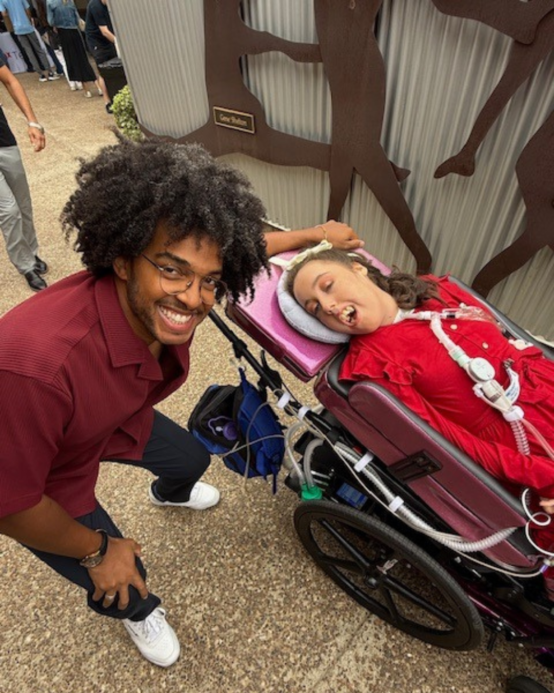 Man kneeling and smiling beside Kennedy reclined in a purple wheelchair with ventilator tubing, outdoors.