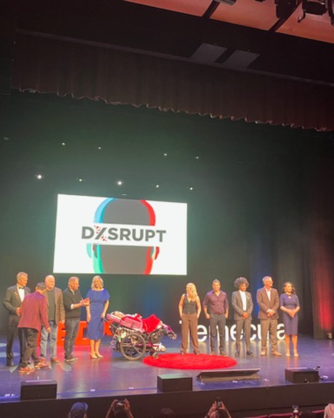 Speakers lined up on a TEDx stage beneath a “DISRUPT” screen, with a young woman in a purple wheelchair near the red carpet.
