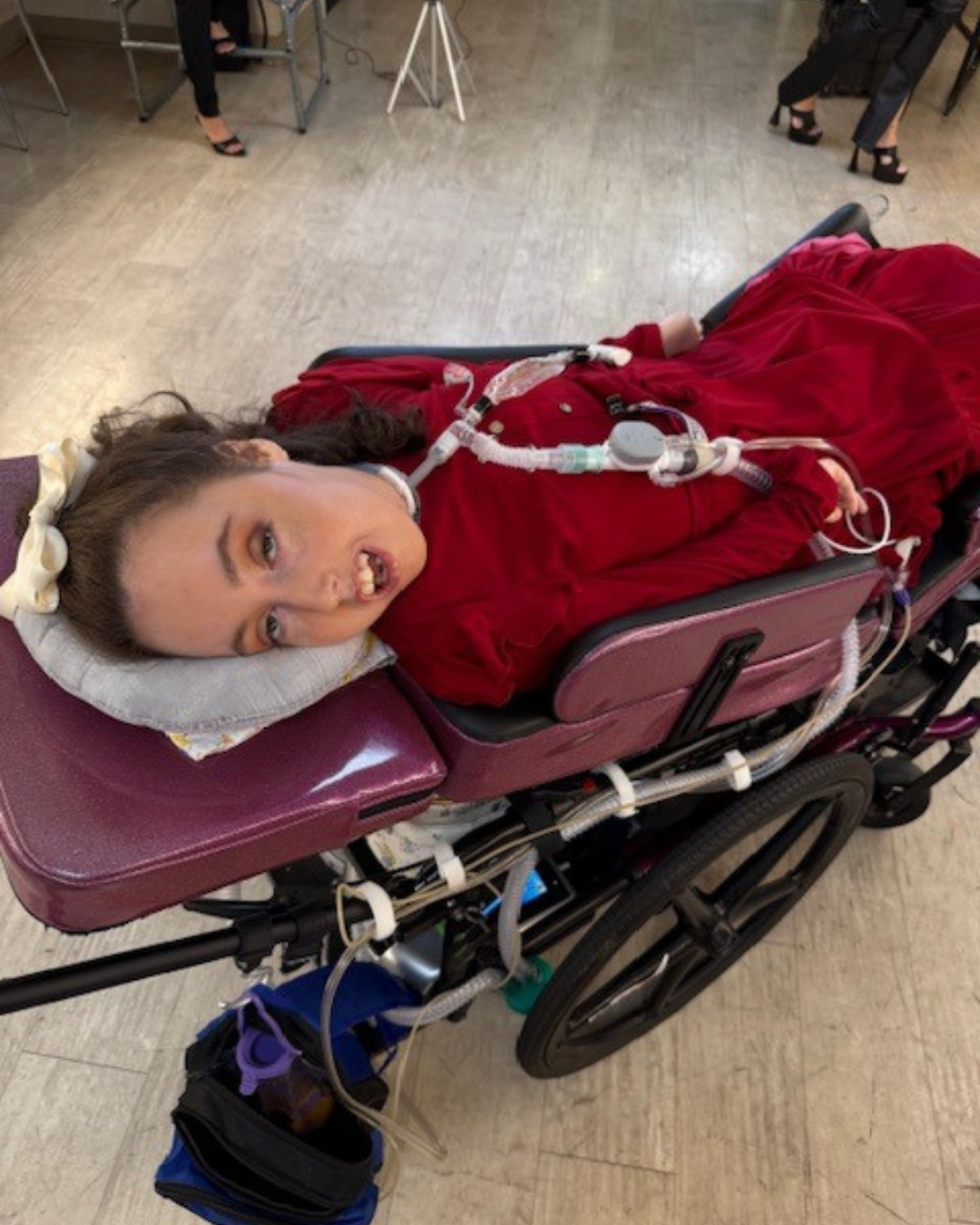 Kennedy reclined in a purple wheelchair wearing a red dress, with ventilator tubing, looking toward the camera indoors.